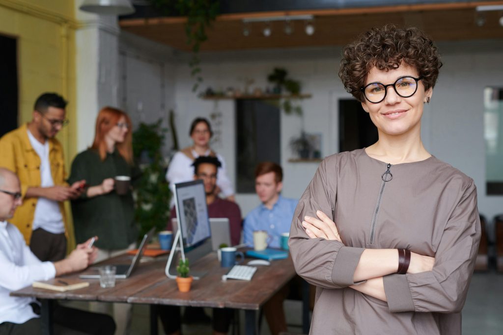 pexels-photo-3184405-3184405 Smiling businesswoman with curly hair stands confidently in a modern office space with colleagues.